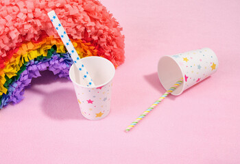 Paper festive cups with pictures of stars and decorative pinata rainbow on a pink background.