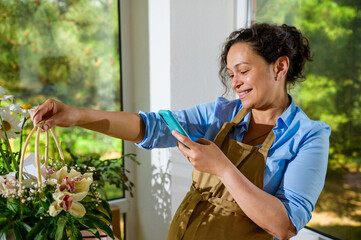 Smiling pregnant woman florist photographing a bouquet of orchid flowers in a wicker basket,...