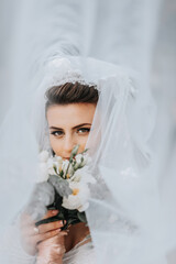 Fototapeta premium The bride poses with a bouquet, covered with a veil. A girl's view through a veil. Wedding dress. Wedding portrait