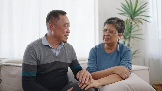 Asian elderly man caring wife holding hands supporting giving sympathy loving while sitting on comfortable sofa in living room at home. Couple retirement lifestyle