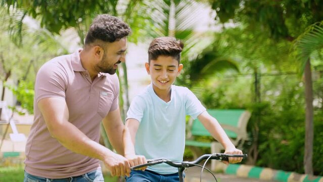 Tracking shot of happy indian father helping his son to ride bicycle at park - concept of Childhood Milestones, Parental Guidance and memories