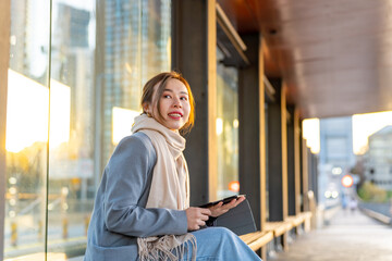 Asian woman freelancer working on digital tablet during waiting for bus at bus stop in the city. Digital nomad people working corporate business from anywhere on gadget device and wireless technology.