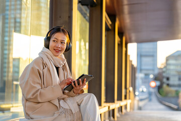 Asian woman listening to the music from headphones and working on digital tablet during waiting for bus in the city. Attractive girl enjoy urban outdoor lifestyle travel with internet on gadget device