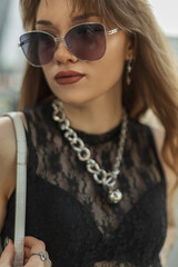 Fashion female street portrait of beautiful stylish hipster woman with cool vintage sunglasses and jewelry in lace black fashion top with bra walks in the city