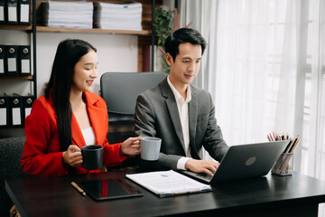 Two Asian business workers talking on the smartphone and using laptop while sitting in office room.