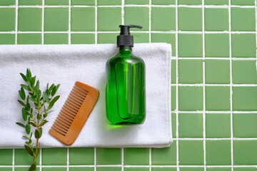 Over the mosaic tiles background, a white towel with a branch of fresh leaves, a wooden comb and an empty label green bottle are displayed. Copy space in the right side