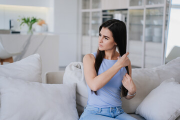 Disappointed brunette hispanic young woman sitting on couch touching hair looks away feels loneliness. Upset caucasian girl at living room combs hair. Crisis, failure. Lady after divorce.