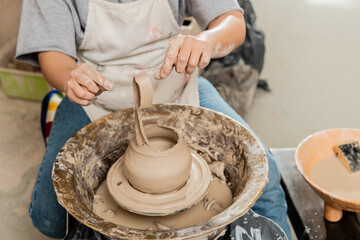Cropped view of fmale artisan in apron holding clay while making vase on spinning pottery wheel near blurred bowl with water in ceramic workshop, artisanal pottery production and process