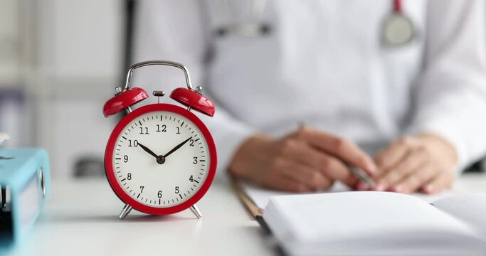 Red alarm clock standing on table in front of doctor closeup 4k movie slow motion. Doctor working day schedule concept