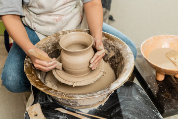 Cropped view of young female potter in apron making shape of clay vase with wooden tool on pottery wheel near bowl with water and sponge in art studio, clay shaping and forming process