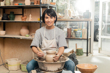 Smiling young brunette asian female artisan in apron looking at camera while holding wooden tool...
