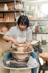 Young asian brunette female artisan in apron and workwear making clay vase and working on spinning pottery wheel in blurred ceramic workshop at background, pottery creation process