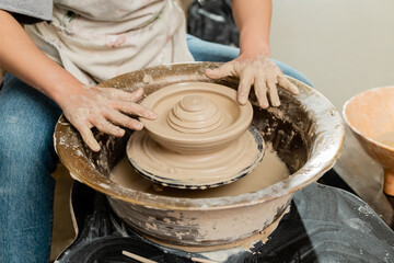 Cropped view of young craftswoman in apron making shape of clay on spinning pottery wheel in blurred ceramic workshop at background, skilled pottery making concept