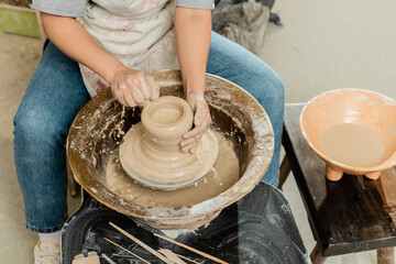 High angle view of young female potter in apron pouring water on clay and spinning pottery wheel near bowl and tools in art workshop, skilled pottery making concept