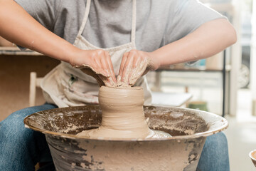 Cropped view of young female potter in apron molding wet clay and working with pottery wheel in blurred art ceramic studio at background, clay sculpting process concept