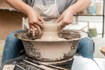 Cropped view of blurred female artisan in apron shaping wet clay on pottery wheel near tools on table in ceramic art workshop ay background, clay sculpting process concept - Powered by Adobe