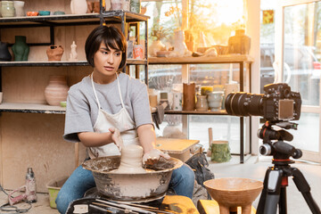 Young asian female artisan in apron shaping wet clay on pottery wheel and talking to digital camera on tripod in blurred ceramic workshop at background, artisan creating unique pottery pieces