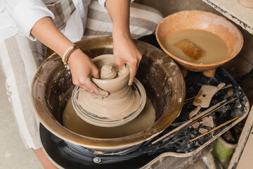 Top view of young female artisan molding wet clay on pottery wheel near tools and bowl with water in ceramic workshop, pottery studio workspace and craft concept