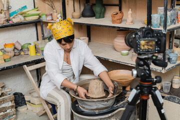 Young asian female artisan in workwear and headscarf molding clay on pottery wheel near blurred...
