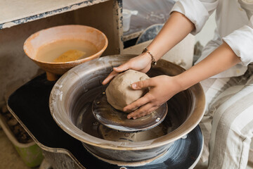 Fototapeta premium Cropped view of young female artisan in workwear molding clay and working with pottery wheel near bowl with water in ceramic workshop, craftsmanship in pottery making