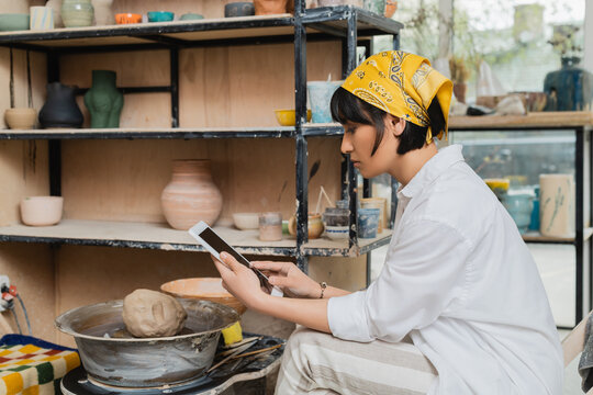 Side View Of Young Brunette Asian Female Artist In Headscarf And Workwear Using Digital Tablet With Blank Screen While Sitting In Ceramic Studio At Background, Craftsmanship In Pottery Making