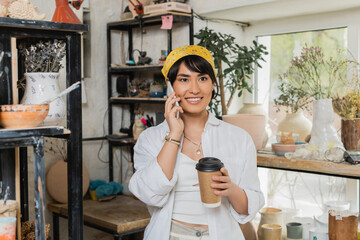 Fototapeta na wymiar Cheerful young asian female potter in headscarf and workwear talking on smartphone and holding coffee to go and standing in blurred pottery workshop, artisan in pottery studio focusing on creation