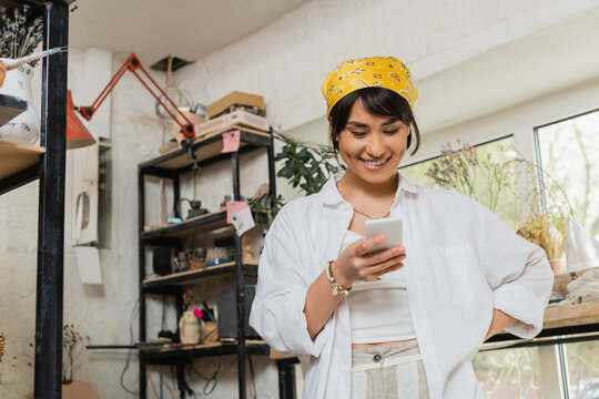 Positive young asian female potter in headscarf and workwear using smartphone while standing and working in blurred ceramic workshop, artisan in pottery studio focusing on creation - Powered by Adobe