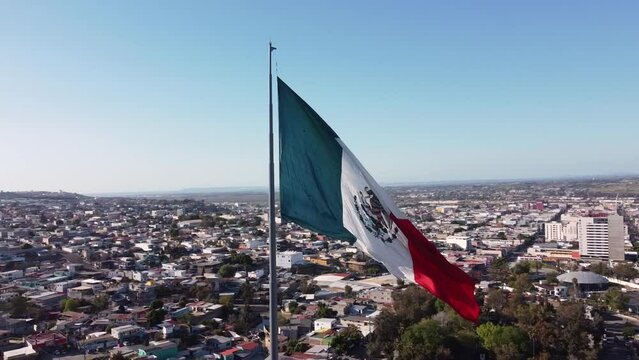 Aerial drone shot of a majestic Mexican flag as it waves over the city of Tijuana (Baja California, Mexico). The camera rotates gently around the flag.