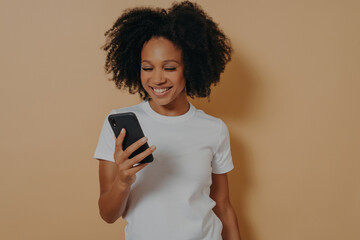 Happy african female student using mobile phone while standing isolated over sand color background