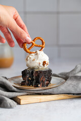 Dark chocolate brownies with scoop of ice cream and salted caramel sauce and pretzels on a grey stone background. Dessert from American cuisine. Woman decorates dessert with pretzel.