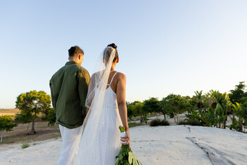 Rear view of newlywed biracial young couple walking at sandy beach amidst trees against clear sky