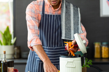 Midsection of biracial man wearing apron preparing meal, composting vegetable waste in kitchen