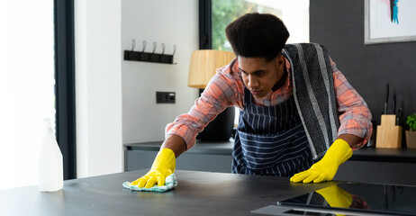 Focused biracial man wearing apron and rubber gloves cleaning countertop in sunny kitchen