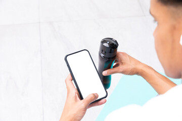 Biracial man using smartphone with copy space, sitting on yoga mat on sunny balcony with water