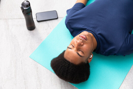 Biracial man doing yoga lying on mat meditating, using smartphone and earphones