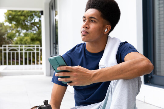Thoughtful biracial man using smartphone and earphones sitting on sunny balcony after yoga