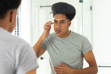 Fototapeta premium Biracial man looking in mirror inspecting face and eyes in sunny bathroom