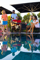 Happy diverse group of friends having pool party and playing guitar in garden
