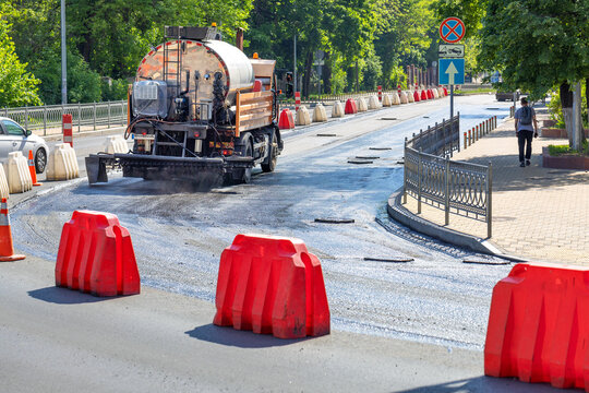 An industrial truck sprays liquid bitumen on a repaired section of a city road before laying new asphalt on a sunny day.