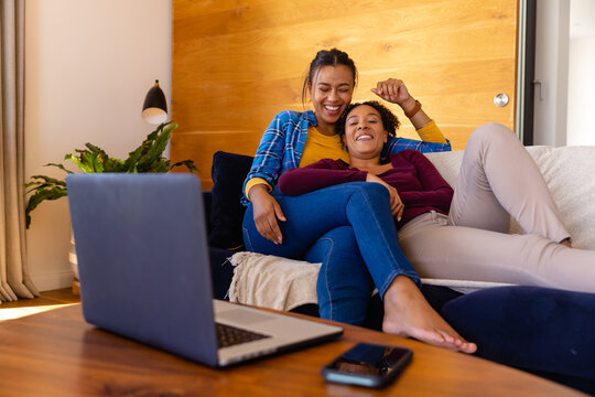 Happy biracial lesbian couple lying on sofa and using laptop in living room
