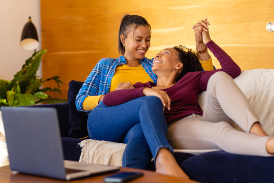 Happy biracial lesbian couple lying on sofa embracing and using laptop in living room