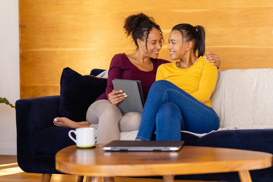 Happy biracial lesbian couple embracing and using tablet on sofa in living room
