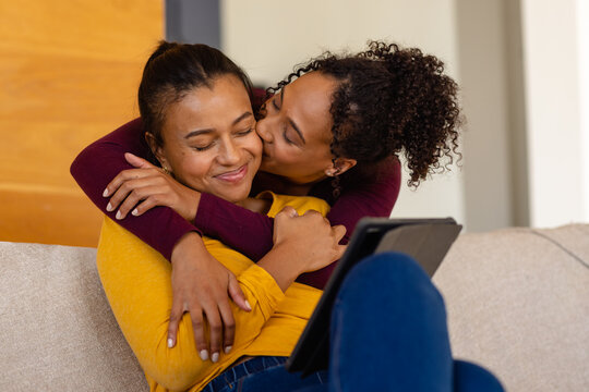 Happy biracial lesbian couple hugging and using tablet on sofa in living room - Powered by Adobe