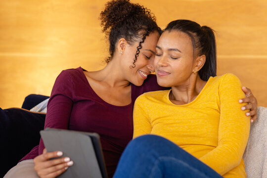 Happy biracial lesbian couple embracing and using tablet on sofa in living room