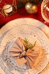 Furoshiki technique. Gift packed in beige fabric, blank card and festive decor on red table, flat lay