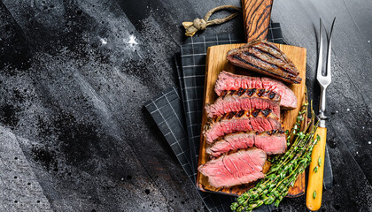 Sliced grilled roast beef with fork for meat on wooden cutting board. Black background. Top view.