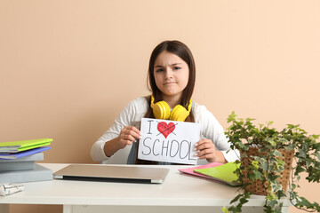 Little girl holding paper with text I DO NOT LOVE SCHOOL at table on beige background