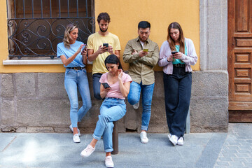 Naklejka na ściany i meble Five young, multiracial friends in urban attire, leaning on a city wall, absorbed in their mobile phones, oblivious to their surroundings.