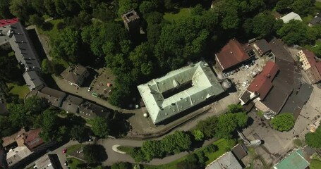 A building of an old brewery next to a park with a historic tower and a large number of green trees during summer in Poland, captured in a 4K bird's-eye view drone shot.
