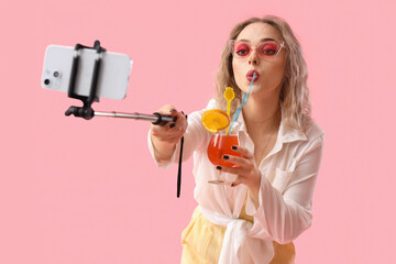 Young woman with summer cocktail and mobile phone taking selfie on pink background